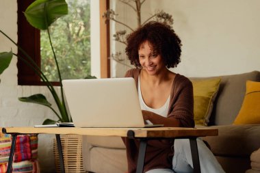 Kendine güveni tam, rahat giyimli genç bir kadın evdeki oturma odasında dizüstünde çalışıyor.