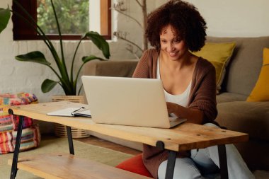 Evde, oturma odasında dizüstü bilgisayarda yazan sıradan kıyafetlerle mutlu bir çift ırklı genç kadın.