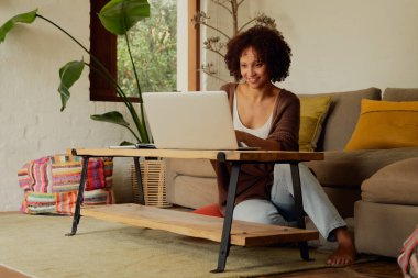 Evde, oturma odasında dizüstü bilgisayarla çalışan, günlük kıyafetli melez genç bir kadın.