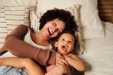 Mutlu çift ırklı anne, günlük giysiler içinde kızını gıdıklıyor ve evde yatakta gülüyor.