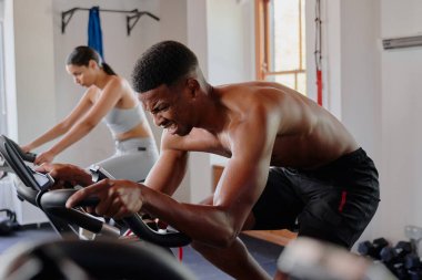 Spor giyim sektöründe, spor salonunda spor bisikleti kullanan kararlı siyahi erkek ve çift ırklı bir kadın.