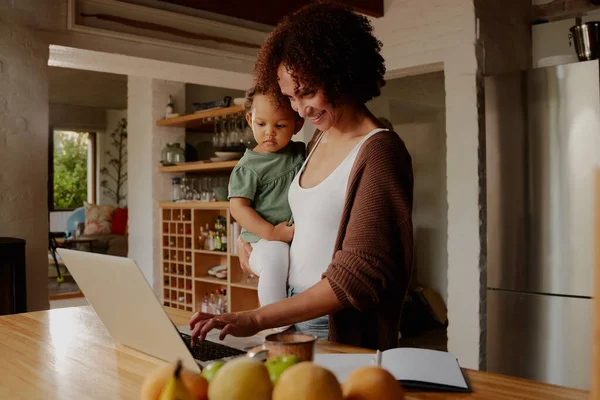 Biracial mother carrying daughter in casual clothing while typing on laptop in kitchen at home