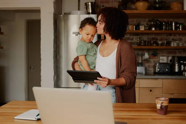 Biracial mother kissing daughter in casual clothing while holding digital tablet by laptop in kitchen at home