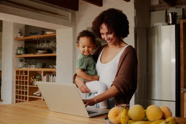 Biracial mother carrying daughter in casual clothing while using laptop in kitchen at home