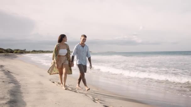 Uczuciowa Młoda Para Uśmiechnięta Trzymająca Się Ręce Spacerując Plaży — Wideo stockowe