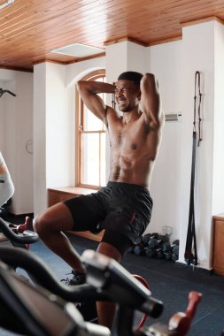 Elleri başının arkasında olan, spor salonunda egzersiz bisikletinde yoğun bir kardiyo çalışması yapan, gömleksiz genç bir zenci.