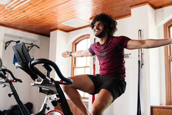 Happy young middle eastern man in sportswear with arms outstretched on exercise bike at the gym