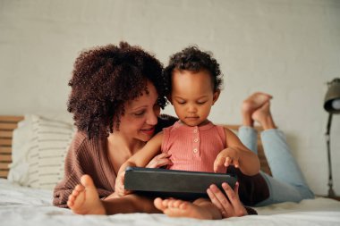Çift ırklı anne ve kız, evde yatakta dinlenirken dijital tablette jest yapıyorlar.