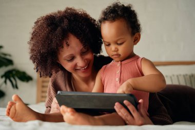 Evde yatakta dijital tablet kullanırken günlük giysiler içinde mutlu anne ve kız.