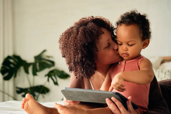 Happy biracial mother kissing daughter on cheek while using digital tablet on bed at home