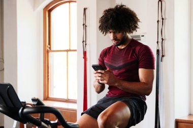 Spor giyim sektöründe odaklanmış genç bir orta doğulu spor salonunda spor bisikletli cep telefonu kullanıyor.