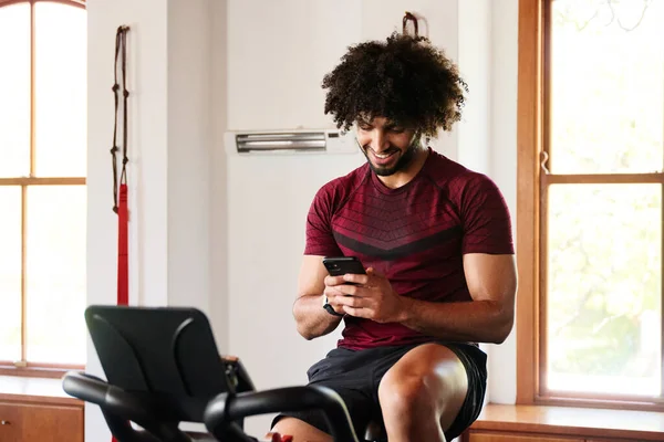 Happy young middle eastern man in sportswear using mobile phone on exercise bike at the gym