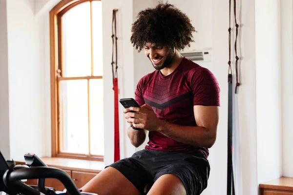 Confident young middle eastern man in sportswear using mobile phone on exercise bike at the gym