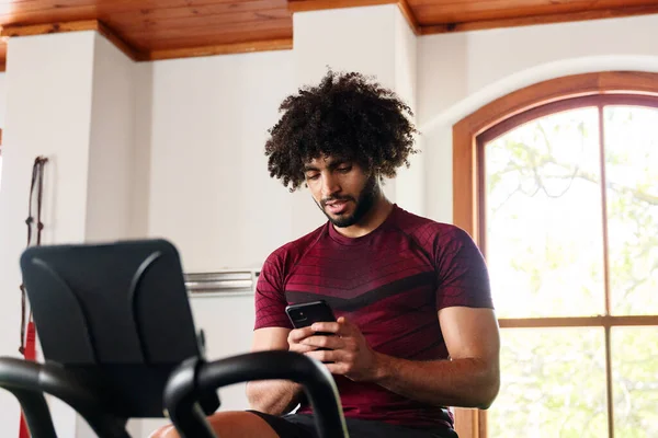 Young middle eastern man in sportswear on mobile phone while using exercise bike at the gym