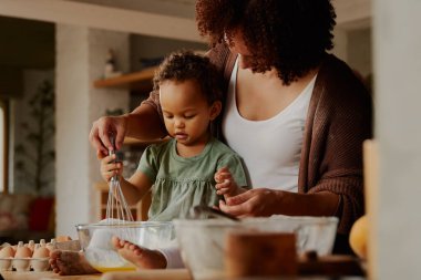 Çift ırklı anne ve kız günlük giysiler içinde evde mutfakta yumurta çırpıyorlar.
