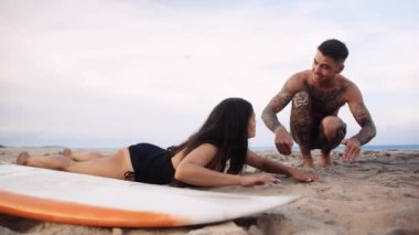 Genç beyaz adam kız arkadaşına mayo öğretiyor. Sahilde sörf tahtası ile kürek çekmeyi öğretiyor.