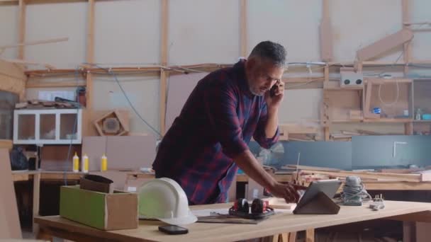 木工工場で携帯電話で話をしながら デジタルタブレット上でスクロールする多人種中大人の男 — ストック動画
