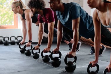 Spor giyim kuşamında bir grup dirençli genç yetişkin spor salonunda kettlebell ile kalas egzersizi yapıyor.