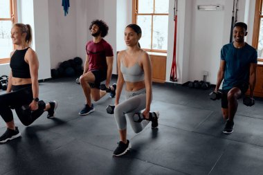 Spor kıyafetleri içinde spor salonunda ağırlıklarla hareket eden bir grup çok ırklı genç yetişkin.