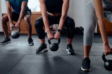 Spor kıyafetleriyle spor salonunda kettlebell çömelmesi yapan üç kararlı genç yetişkin.