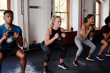 Spor giyim kuşamında, spor salonunda arka arkaya kettlebell çömelmesi yapan bir grup esnek, çok ırklı genç yetişkin.