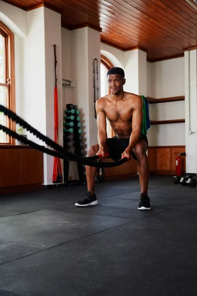 Shirtless young black man sweating while doing battle ropes exercises at the gym