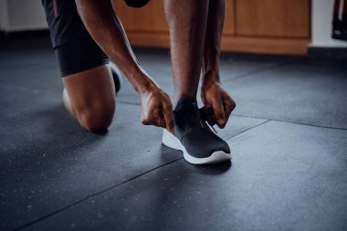 Spor salonunda diz çöküp ayakkabı bağlayan genç siyahi adamın yakın çekimi.