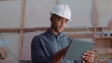 Ahşap fabrikasında çalışırken dijital tablet kullanan güvenlik kasklı çok ırklı genç adam.