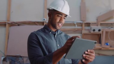 Marangozluk fabrikasında çalışırken dijital tablete kayan kasklı çok ırklı genç bir adam.
