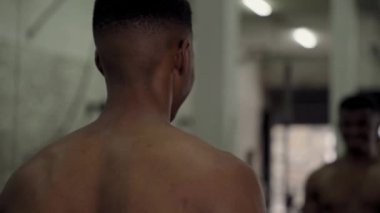 Azimli, üstsüz, kaslı, genç siyahi adam spor salonunda ayna ile halter kaldırıyor.