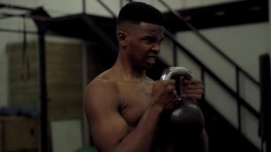 Azimli, kaslı, genç siyah adam spor salonunda kettlebell ile ağırlık kaldırıyor.