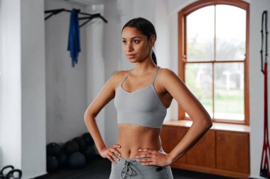 Spor giyim sektöründe odaklanmış, elleri kalçasında spor salonunda başka tarafa bakan iki ırklı bir kadın.