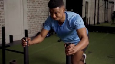 Spor giyimli, kaslı, genç, siyah bir adam spor salonunda kızak itme egzersizi yapıyor.