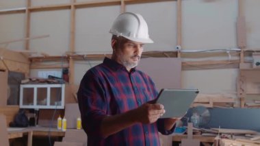 Güvenlik kaskı takan çok ırklı orta yaşlı bir adam marangozluk fabrikasında dijital tablet üzerinde çalışıyor.