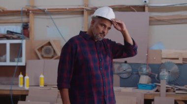 Çok ırklı, profesyonel, orta yaşlı bir adam marangozluk fabrikasında çalışırken kaskını çıkarıyor.