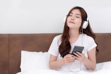 Güzel Asyalı kadınlar kulaklık takıp akıllı telefonlarından müzik dinliyorlar. Beyaz yatak odasındaki yatakta.