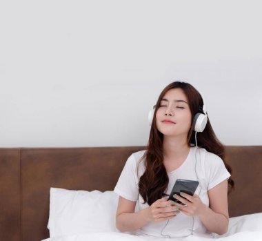 Güzel Asyalı kadınlar kulaklık takıp akıllı telefonlarından müzik dinliyorlar. Beyaz yatak odasındaki yatakta.