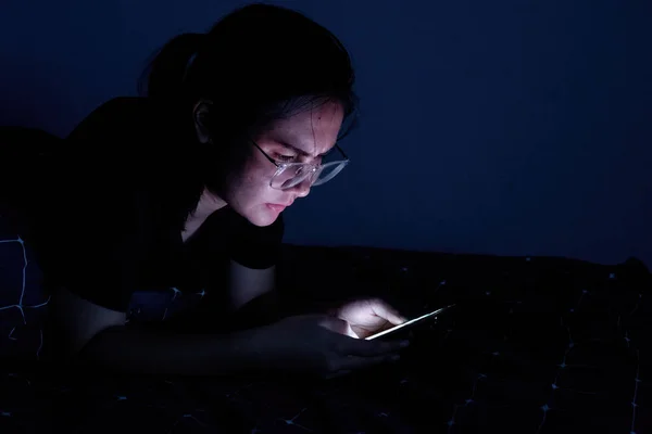 亚洲女人戴眼镜是在用一个屏幕非常明亮的智能手机 弱光下蓝光的危险 — 图库照片