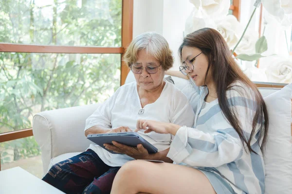 Asyalı bir kadın, annesine yaşlı bakım konseptinde tablet kullanmayı öğretiyor..