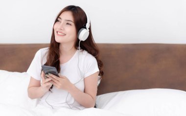 Güzel Asyalı kadınlar kulaklık takıp akıllı telefonlarından müzik dinliyorlar. Beyaz yatak odasındaki yatakta.