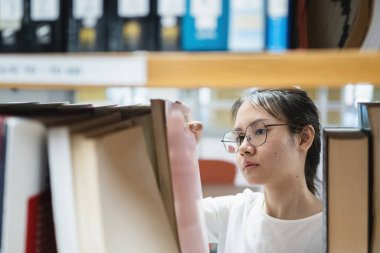 Eğitim ve kütüphane kavramı, gözlüklü Asyalı bir kadın kitaplıktan kitap seçiyor..