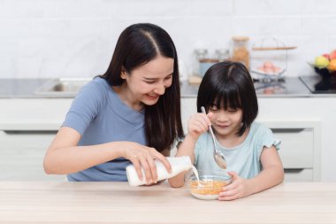 Güzel bir kadın ve tatlı bir kız Asyalılar süt ve mısır gevreği yiyorlar. Evdeki mutfak masasında mutlu bir kahvaltı. Bu aile için yeni bir normal hayat..