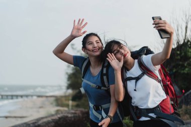 Outdoor tourism concept, two beautiful Asian backpackers taking selfies and video calls using smartphones.