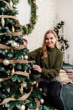 Sarışın, güzel bir genç kadın bir Noel ağacının yanında oturup oyuncaklarla süslüyor. Yeni yıl yakında. Sıcak bir evde Noel atmosferi