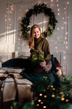 Noel için dekore edilmiş bir yatak odasında süveter giyen genç bir kadın Noel ağacının zeminine karşı Noel hediyelerini açıyor..