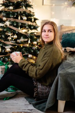 Sarışın, güzel genç bir kadın elinde kahveyle bir Noel ağacının yanında oturuyor. Yeni yıl yaklaşıyor. Sıcak bir evde Noel atmosferi