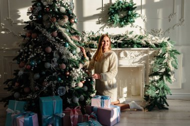 Sarışın, güzel bir genç kadın bir Noel ağacının yanında oturup oyuncaklarla süslüyor. Yeni yıl yakında. Sıcak bir evde Noel atmosferi