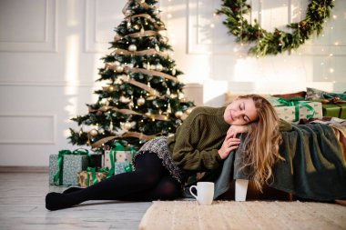 Sarışın, güzel genç bir kadın elinde kahveyle bir Noel ağacının yanında oturuyor. Yeni yıl yaklaşıyor. Sıcak bir evde Noel atmosferi