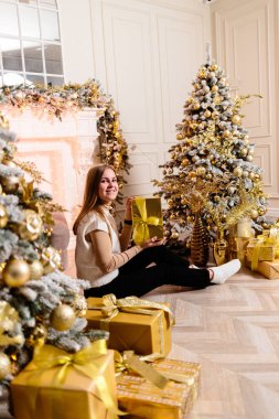 Süveter giyen güzel bir kız çelenklerle süslenmiş bir Noel ağacının yanında oturur. Yanındaki Noel ağacı hediye kutularıyla süslenmiş. Yeni Yıl