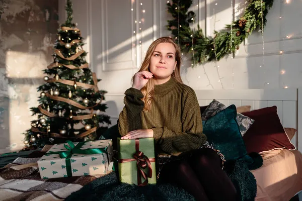 一位身穿毛衣的年轻女子在圣诞树旁装饰着圣诞装饰品的卧室里打开圣诞礼物 — 图库照片